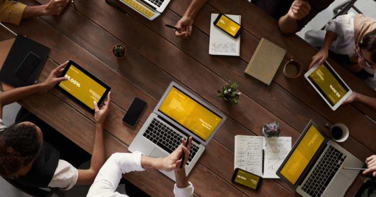 An overhead view of a business meeting, featuring laptops, tablets, and various people around a wooden table.